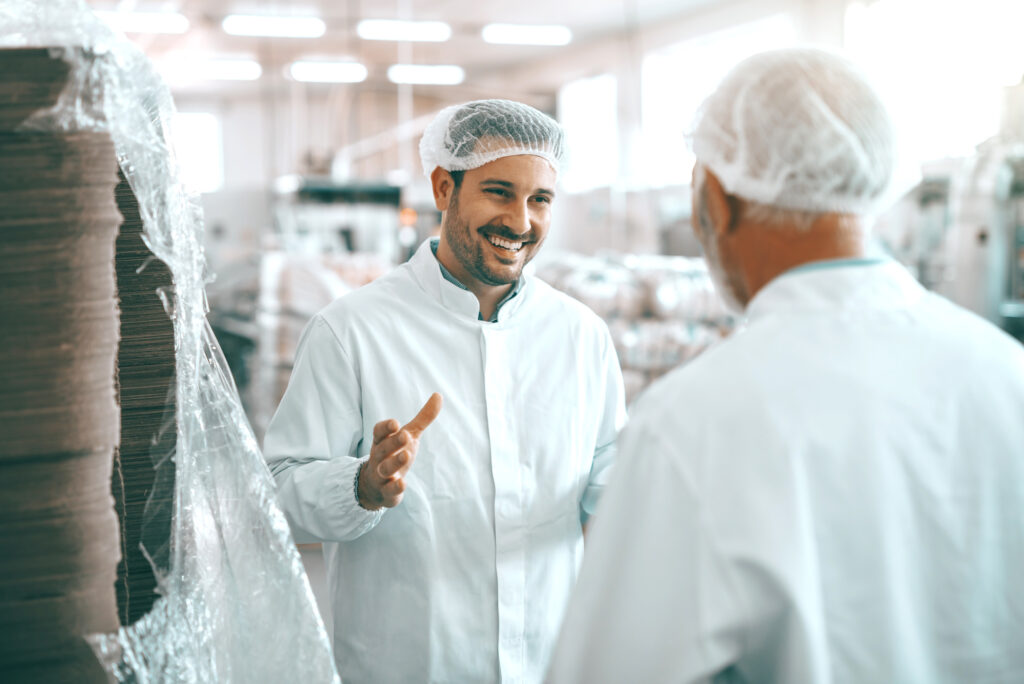 Two cheerful Caucasian food plant workers in white uniforms and with hairnets standing and talking about food production.